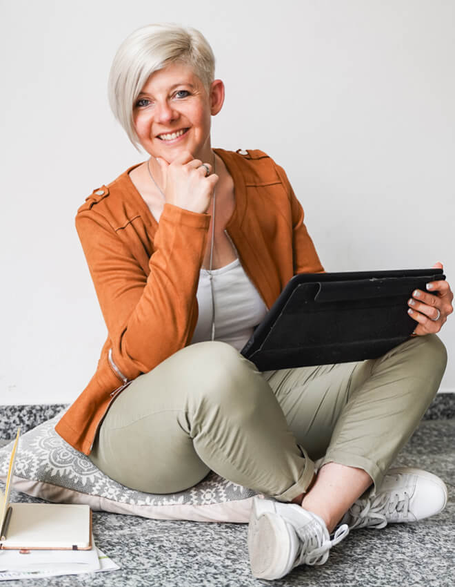 Frau (Julia) mit Tablet sitzt lächelnd auf einem Bodenkissen, Notizbuch liegt daneben.