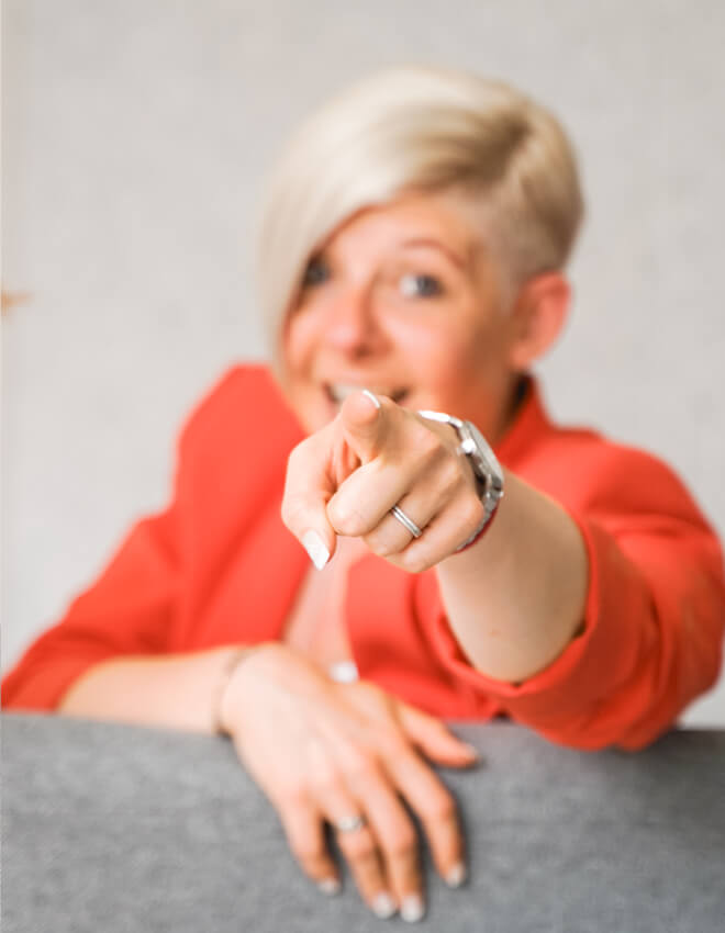 Frau (Julia) mit blondem Kurzhaarschnitt zeigt direkt in die Kamera und lächelt.