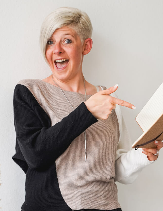 Fröhliche Frau (Julia) mit Notizbuch zeigt lachend darauf und blickt in die Kamera.