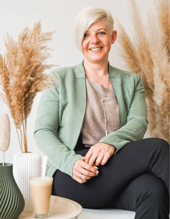 Julia mit blondem Kurzhaarschnitt sitzt lächelnd vor Trockenblumen-Deko mit Latte Macchiato auf dem Tisch.