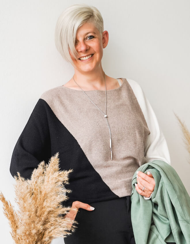 Lächelnde Frau (Julia) mit asymmetrischem Pullover vor heller Wand, hält grüne Jacke in der Hand.