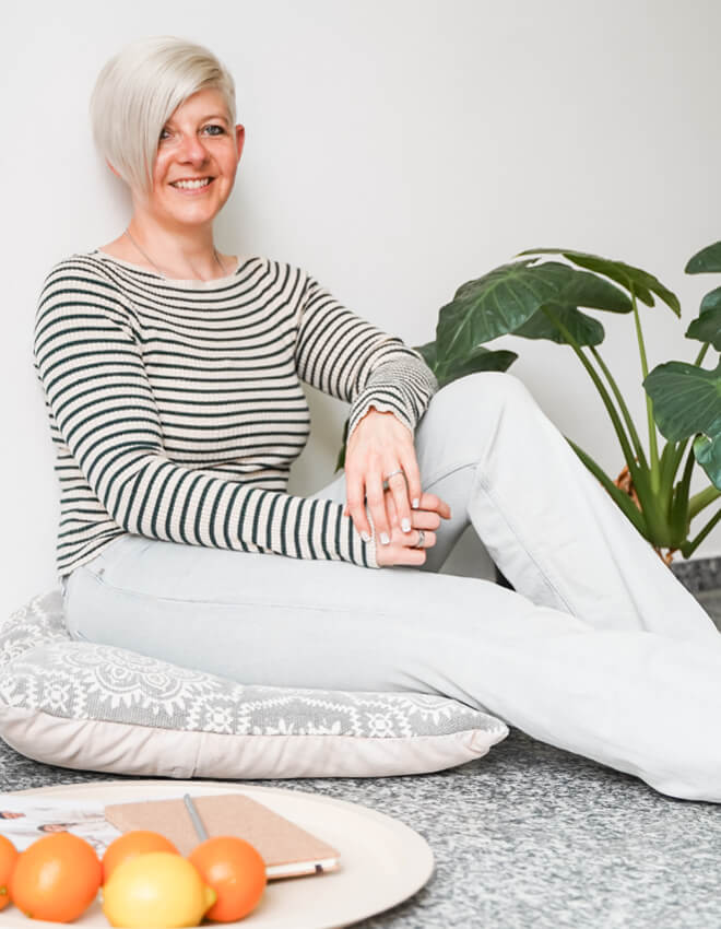 Frau (Julia)mit Kurzhaarfrisur sitzt entspannt auf einem Bodenkissen neben einer Pflanze, im Vordergrund liegt Obst auf einem Tablett.