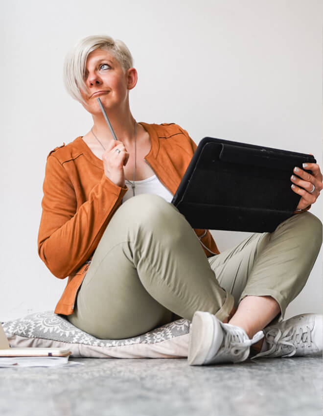 Frau (Julia) sitzt auf einem Kissen mit Tablet und Stift, blickt nachdenklich zur Seite.