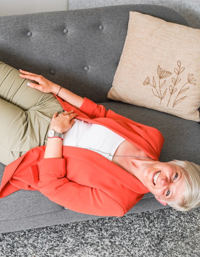 Lächelnde Frau (Julia) liegt entspannt auf einem grauen Sofa mit besticktem Kissen.
